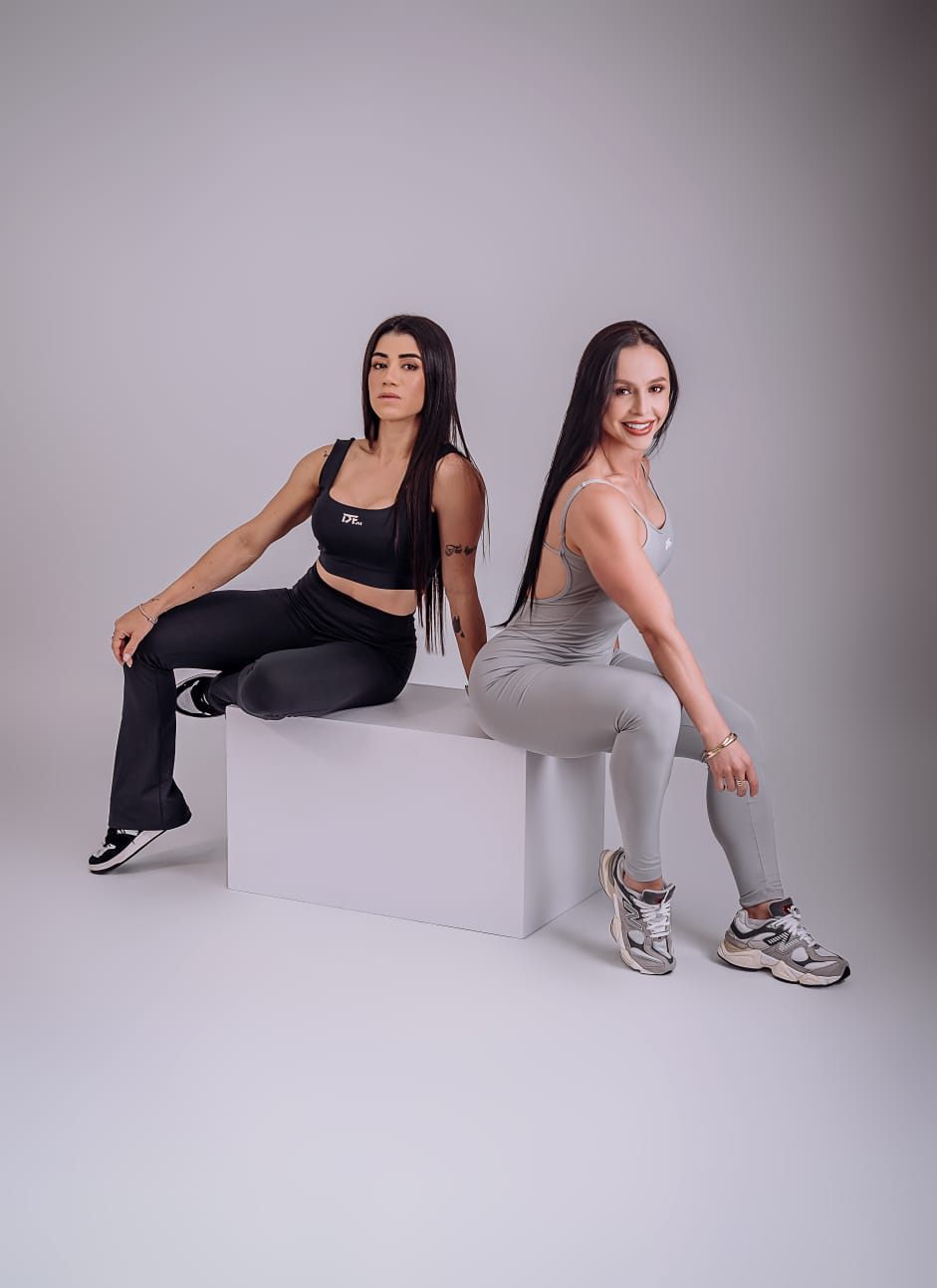 Duas personal trainers sentadas em um cubo branco, em ensaio de estúdio
